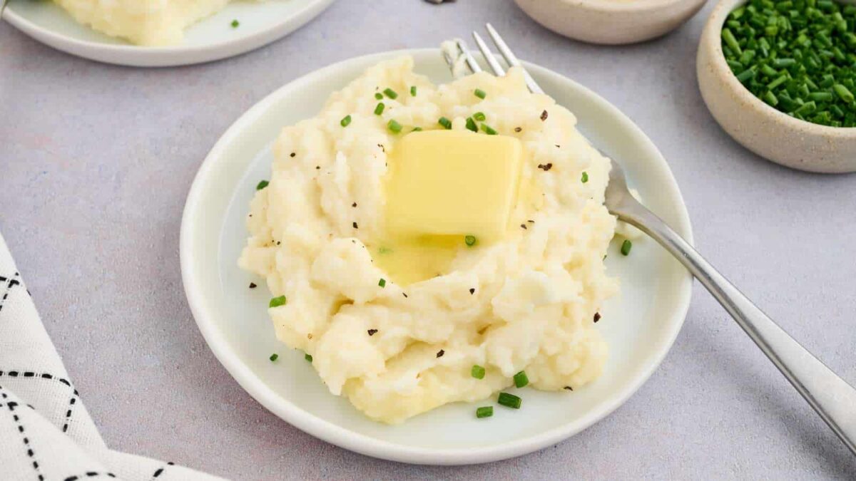 A plate of creamy mashed potatoes topped with a pat of butter and chopped chives, with a fork on the side—classic comforting food.