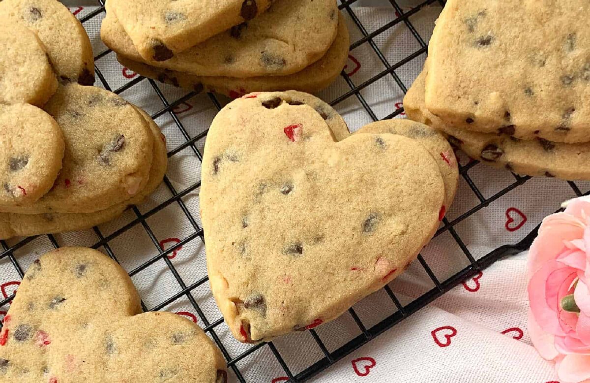 Heart-shaped chocolate chip cookies with red candy pieces cooling on a black wire rack—easy cookies that come together in minutes. A pink flower and heart-patterned cloth complete this sweet, charming scene.