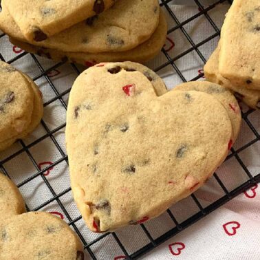 Heart-shaped chocolate chip cookies with red candy pieces cooling on a black wire rack—easy cookies that come together in minutes. A pink flower and heart-patterned cloth complete this sweet, charming scene.