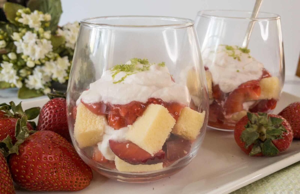 Two glasses filled with layers of sponge cake cubes, sliced strawberries, and whipped cream, garnished with lime zest—these allergy friendly snacks make perfect nut free desserts. Whole strawberries are on the plate next to the glasses.
