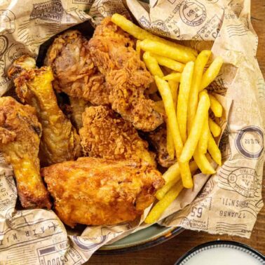 A basket lined with printed paper holds fried chicken pieces and French fries, perfect for easy dinners, placed on a wooden table next to small dishes of spices and salt.