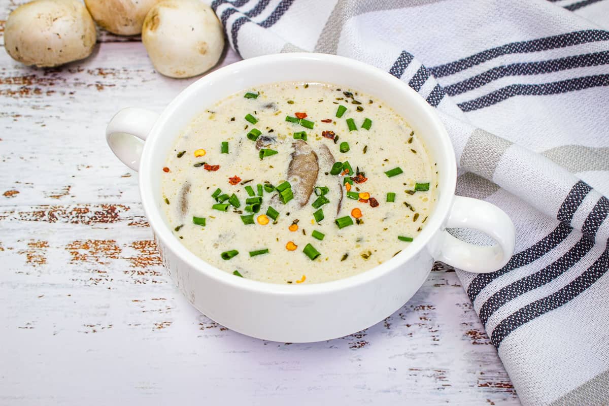 A bowl of creamy mushroom soup—one of the best comfort soups—garnished with chopped chives and red pepper flakes, sits on a rustic white table with a striped cloth and mushrooms in the background.