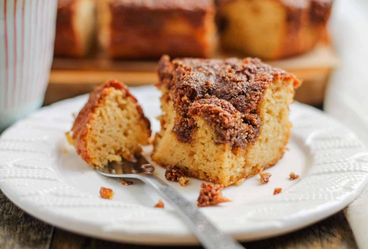 A slice of crumb-topped gluten free coffee cake sits on a white plate with a fork, with more gluten free cakes in the background.