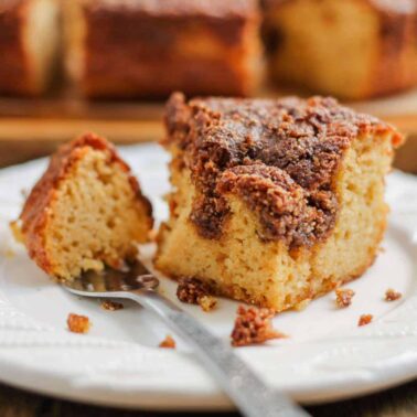 A slice of crumb-topped gluten free coffee cake sits on a white plate with a fork, with more gluten free cakes in the background.