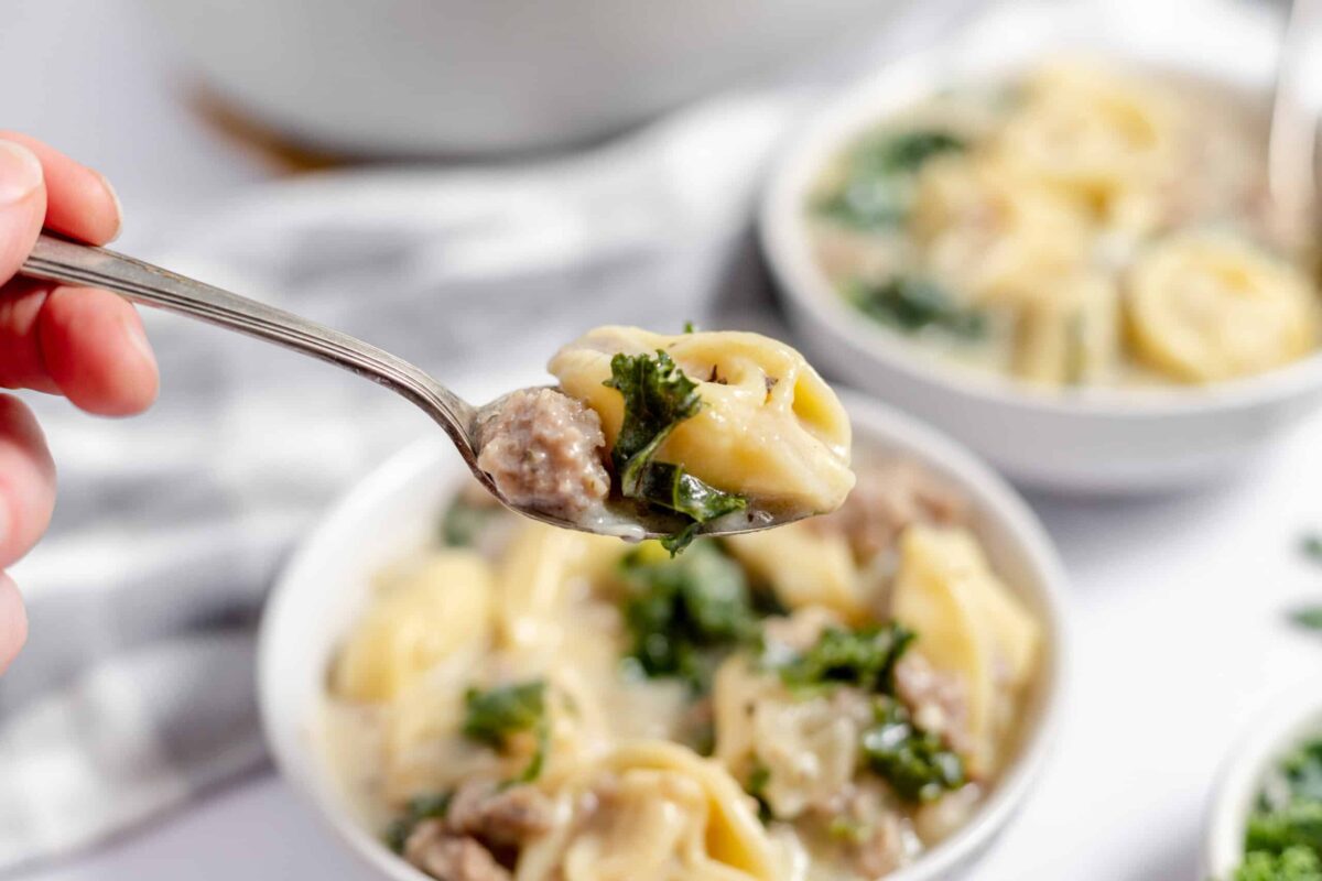 A spoon holding tortellini, sausage, and kale hovers above a bowl of creamy comfort soup—perfect for those who love hearty comfort food and crave new soup recipes.