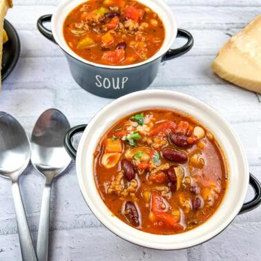 Two bowls of hearty comfort soup with beans, ground meat, and tomatoes sit on a white surface, alongside two spoons, sliced bread, and a wedge of cheese—a perfect scene for those who love comforting soup recipes.