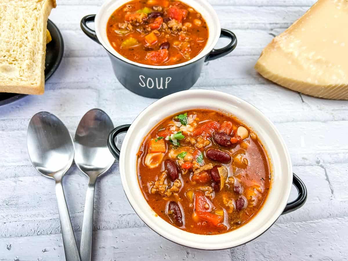 Two bowls of hearty comfort soup with beans, ground meat, and tomatoes sit on a white surface, alongside two spoons, sliced bread, and a wedge of cheese—a perfect scene for those who love comforting soup recipes.
