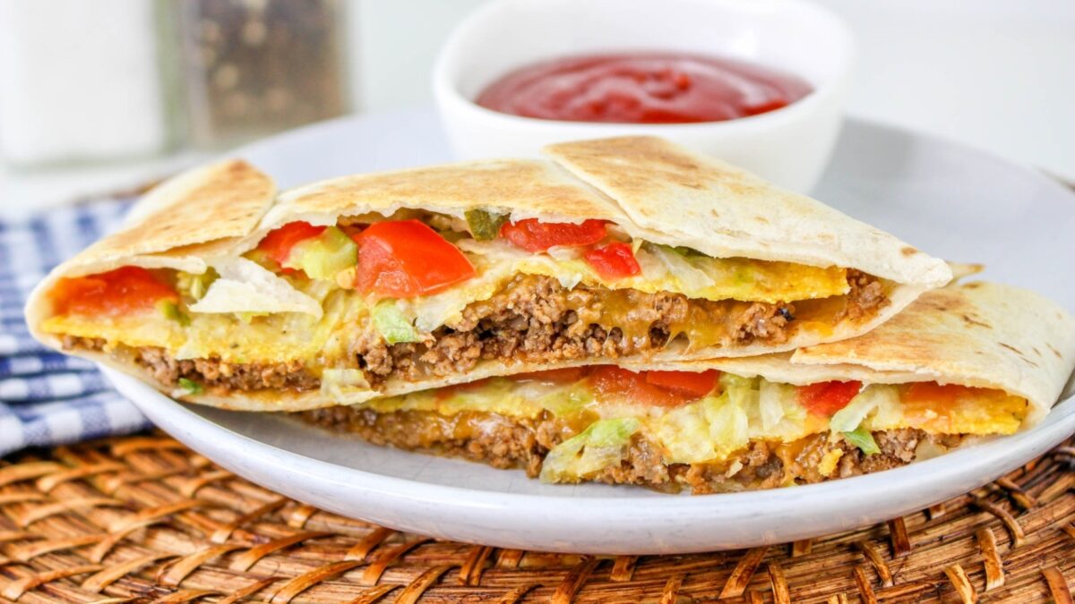 A quesadilla filled with ground beef, lettuce, tomato, and cheese is cut in half and served on a plate with a side of red dipping sauce—perfect for anyone seeking tasty recipes or affordable meals.