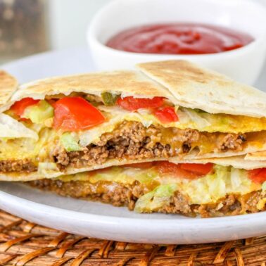 A quesadilla filled with ground beef, lettuce, tomato, and cheese is cut in half and served on a plate with a side of red dipping sauce—perfect for anyone seeking tasty recipes or affordable meals.