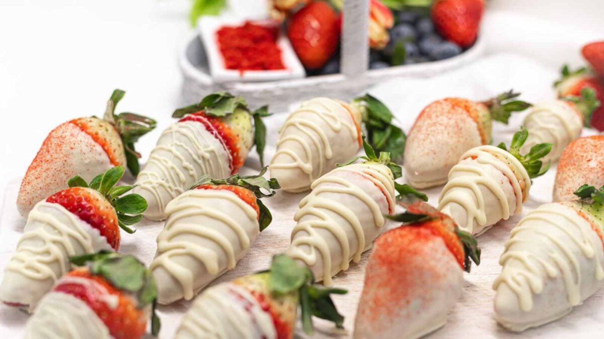 Fresh strawberries coated in white chocolate, some drizzled with more chocolate and others dusted with red powder, evoke inviting aromas of cozy desserts, arranged on a white surface with a basket of berries in the background.