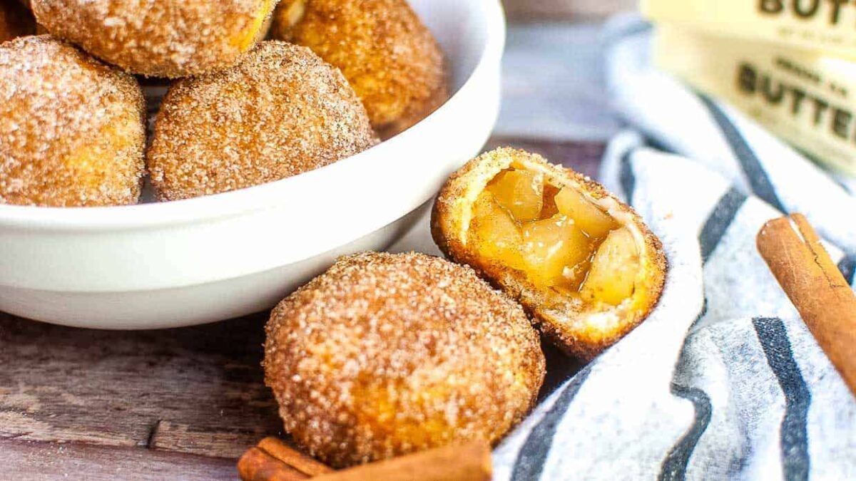 A bowl of sugar-coated dough balls, with one cut open to reveal an apple filling—perfect kid friendly snacks. Two cinnamon sticks and a butter package are nearby on a wooden surface.