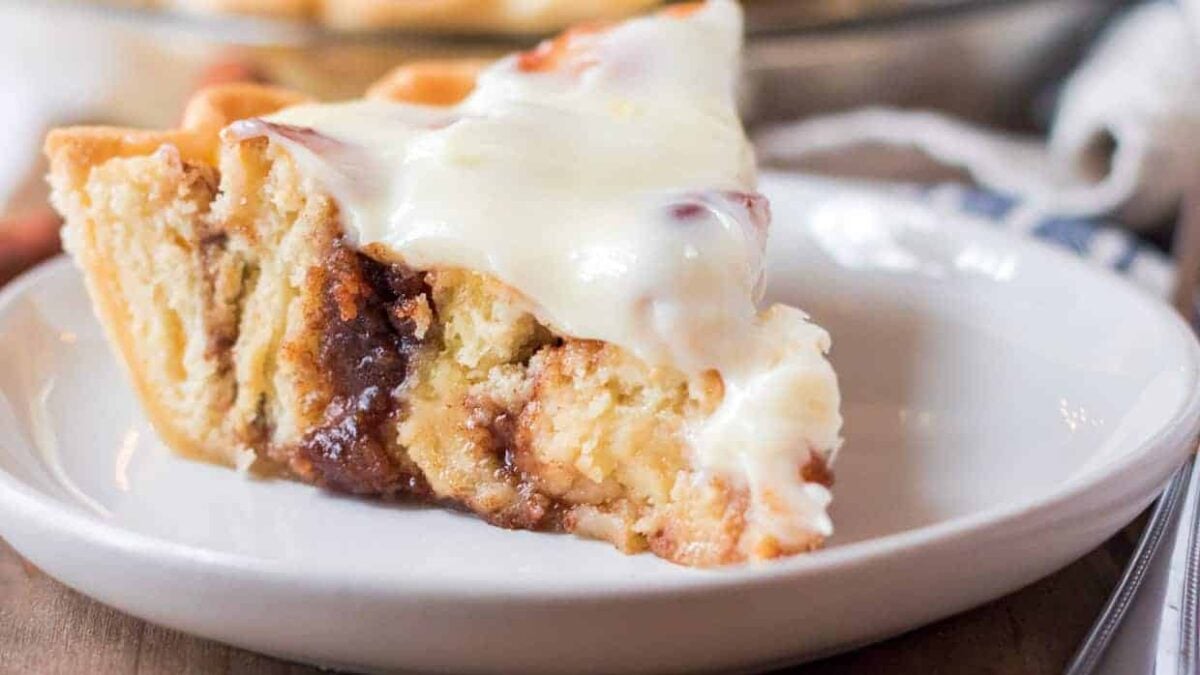 A slice of cinnamon roll pie topped with a thick layer of white icing sits on a white plate, perfect for fans of cozy desserts and inviting aromas.