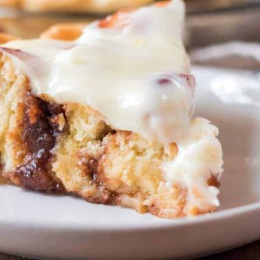 A slice of cinnamon roll pie topped with a thick layer of white icing sits on a white plate, perfect for fans of cozy desserts and inviting aromas.