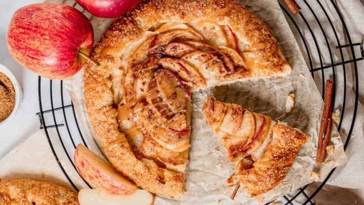 A rustic apple galette with a slice missing sits on parchment paper atop a wire rack, surrounded by fresh apples and cinnamon sticks—perfect among cute fall desserts or as festive Halloween snacks.