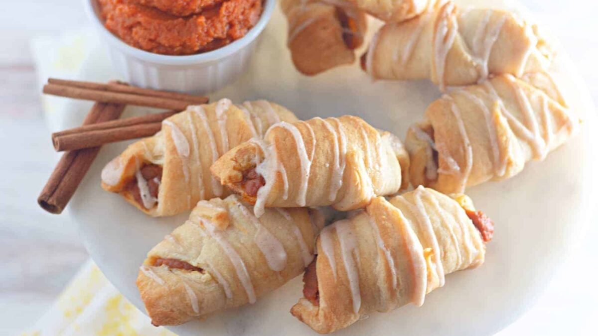 Six crescent rolls with icing drizzle are arranged on a white plate, accompanied by a small bowl of orange spread and two cinnamon sticks—perfect as cute fall desserts or festive Halloween snacks.