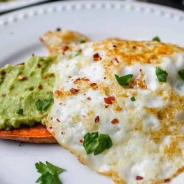 A plate with two fried eggs topped with herbs and red pepper flakes, served alongside mashed avocado on a slice of sweet potato—perfect for those seeking easy breakfast recipes.