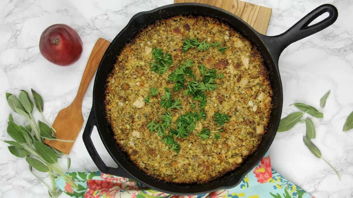 A cast iron skillet filled with baked stuffing topped with fresh parsley, surrounded by a red onion, sage leaves, and a wooden spatula on a marble surface—perfect for Thanksgiving sides or other holiday side dishes.