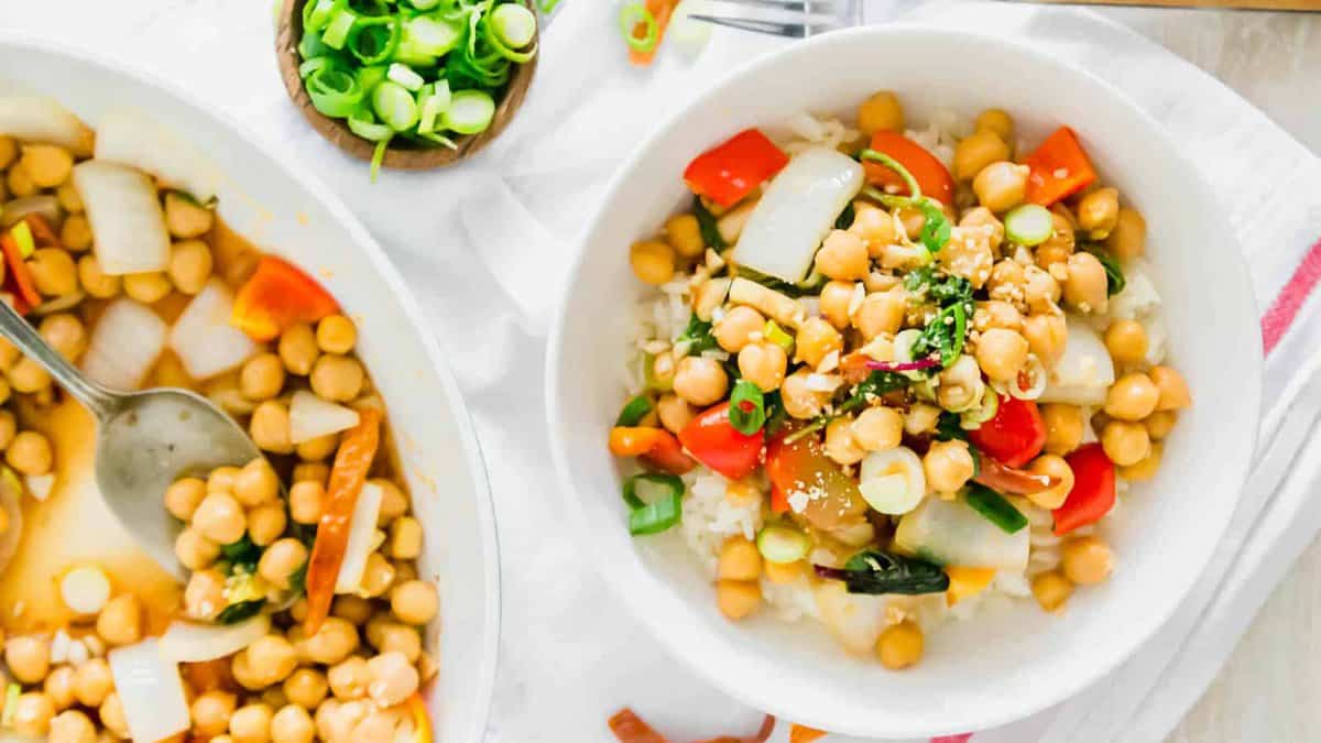A bowl of rice topped with chickpeas, diced red bell peppers, onions, green onions, and sesame seeds—an easy option for vegetarian dinners—with a similar mixture cooking in a pan beside it.