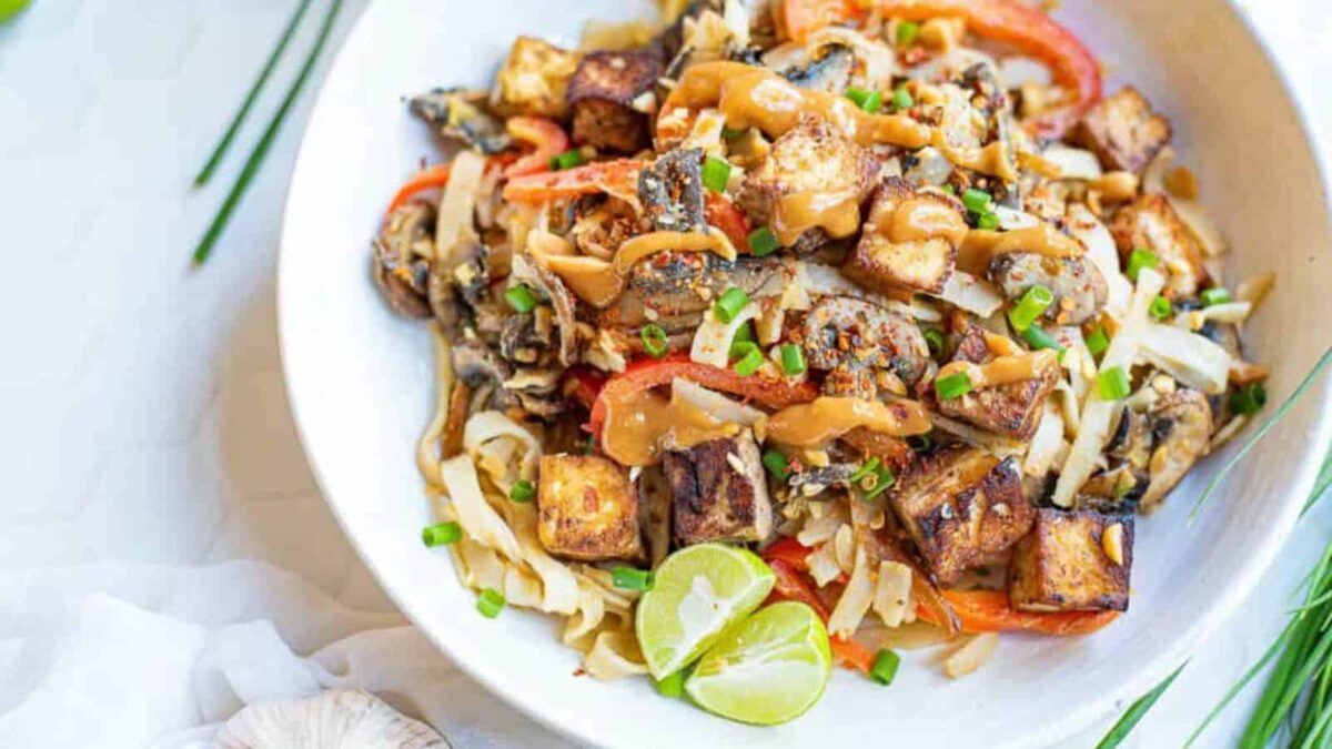 A bowl of noodle stir-fry topped with tofu cubes, sliced vegetables, chopped chives, and a drizzle of sauce, garnished with lime wedges—perfect for those seeking flavorful vegetarian recipes or easy meatless meals.