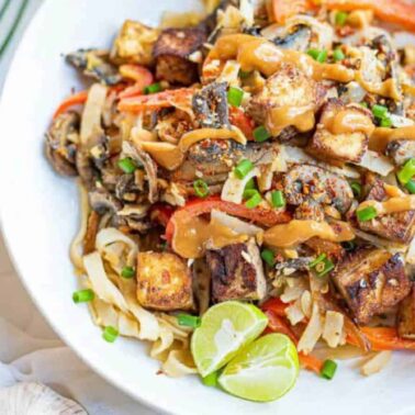 A bowl of noodle stir-fry topped with tofu cubes, sliced vegetables, chopped chives, and a drizzle of sauce, garnished with lime wedges—perfect for those seeking flavorful vegetarian recipes or easy meatless meals.