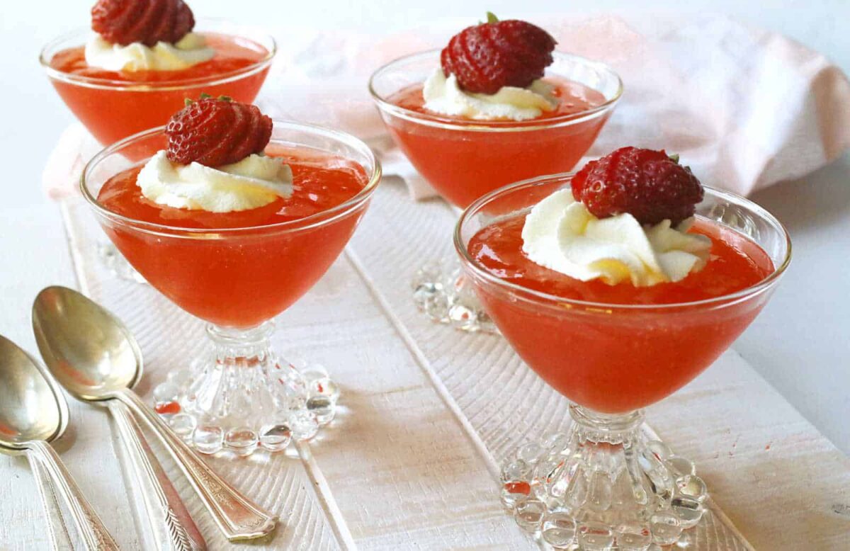 Four glass dessert cups filled with strawberry mousse, each topped with whipped cream and a strawberry, sit beside three silver spoons on a white surface—an irresistible treat for fans of strawberry desserts.