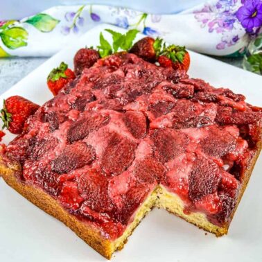 A square strawberry upside-down cake with a piece missing, garnished with fresh strawberries, on a white plate next to a floral napkin and purple flowers—an irresistible addition to any collection of strawberry desserts.