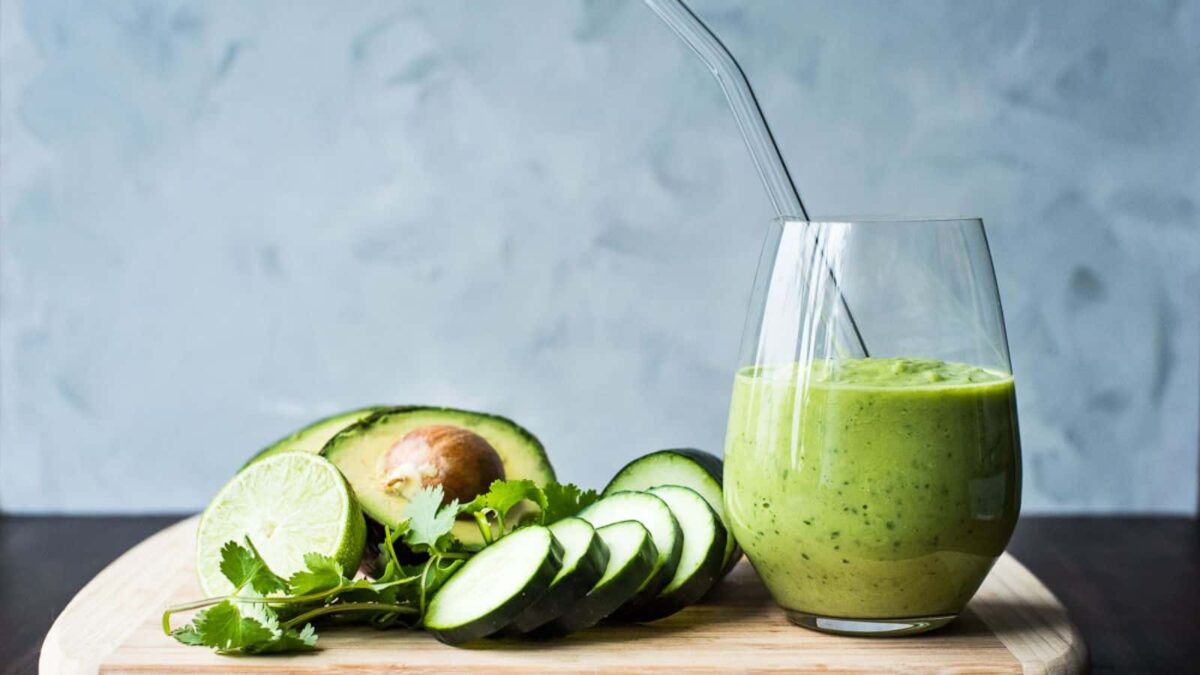 A glass of green smoothie with a straw sits next to sliced cucumber, a halved avocado, lime halves, and cilantro on a wooden surface—perfect inspiration for delicious smoothie recipes and other healthy treats.
