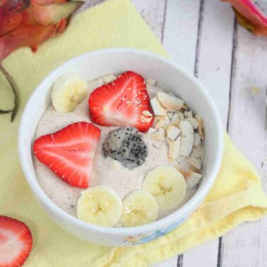 A smoothie bowl, perfect for fans of smoothie recipes, is topped with sliced strawberries, bananas, dragon fruit, and coconut flakes. It sits on a yellow napkin next to a spoon—an irresistible treat for any morning!.
