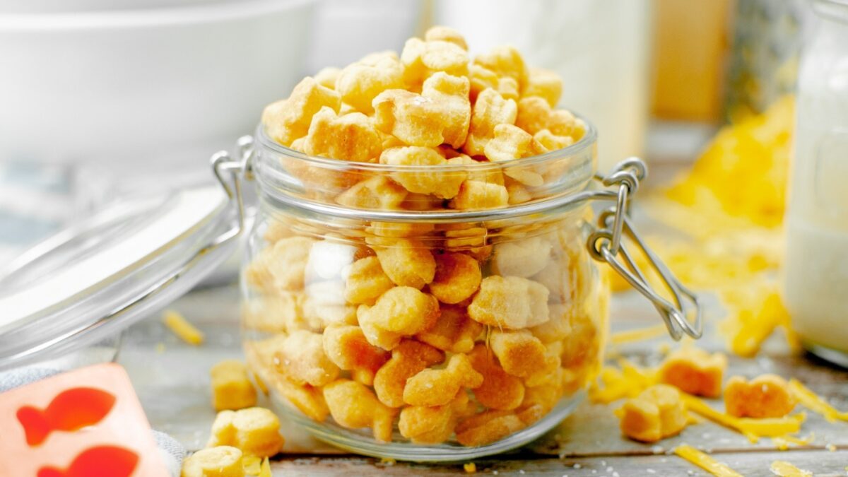 A glass jar filled with small, goldfish-shaped cheese crackers sits on a wooden surface with a few crackers scattered around—perfect kid friendly snacks for any occasion.