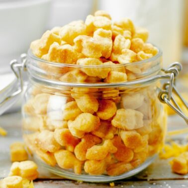 A glass jar filled with small, goldfish-shaped cheese crackers sits on a wooden surface with a few crackers scattered around—perfect kid friendly snacks for any occasion.