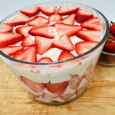 A glass bowl filled with strawberry trifle, featuring layers of sliced strawberries, cream, and cake, sits beside a bowl of whole strawberries on a wooden surface—an irresistible option for fans of no bake desserts and easy strawberry recipes.