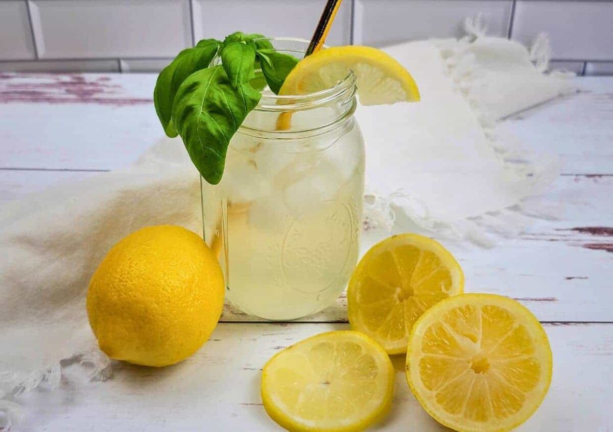 A mason jar filled with iced lemonade, garnished with basil and a lemon wedge, sits on a white surface surrounded by a whole lemon and slices—perfect inspiration for refreshing lemon recipes.