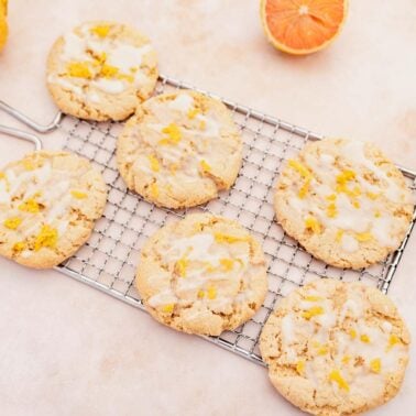 Six glazed citrus cookies are arranged on a cooling rack, surrounded by whole and sliced citrus fruits—a delightful scene for anyone who loves lemon desserts.