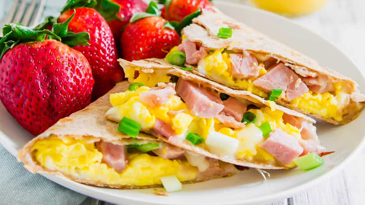 A plate with three ham, scrambled egg, and cheese quesadilla slices, garnished with chopped green onions, sits next to fresh strawberries—perfect for healthy breakfast lovers looking for easy breakfasts.