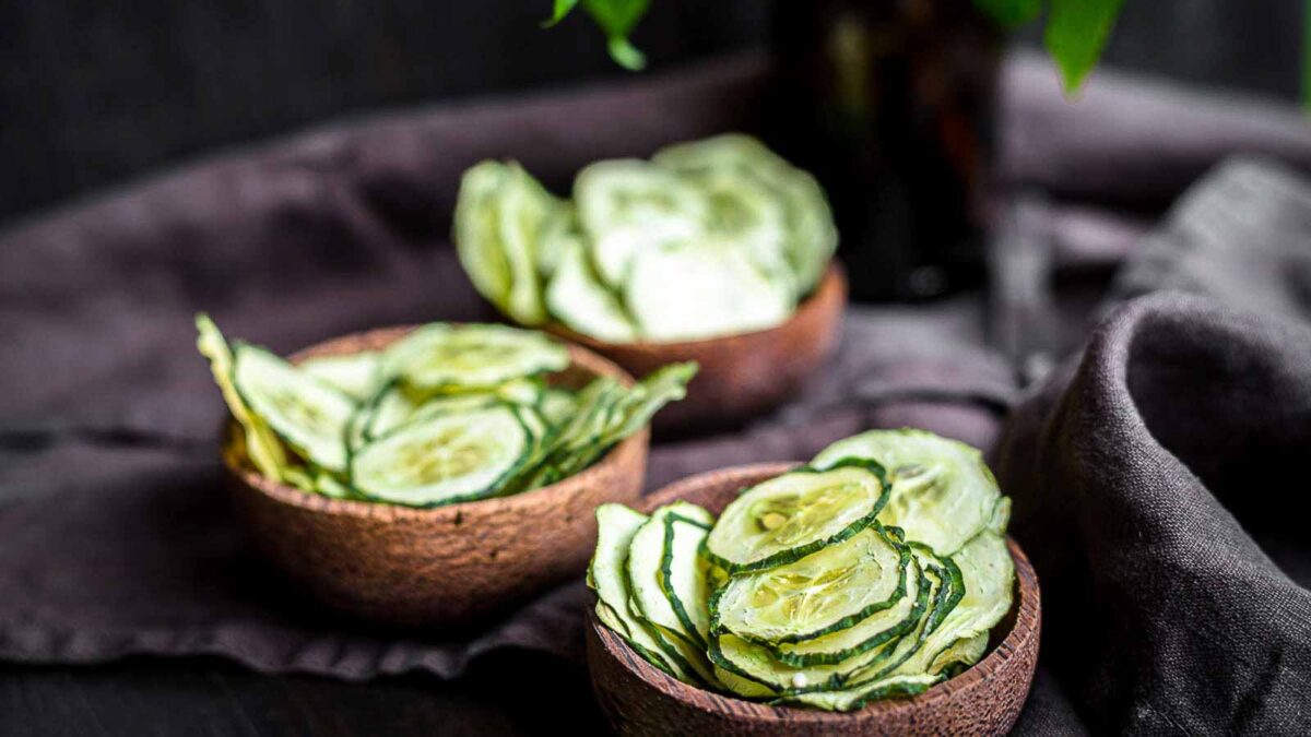 Three wooden bowls filled with thinly sliced cucumber chips rest on a dark cloth, offering fresh and delicious snack ideas against a blurred background.