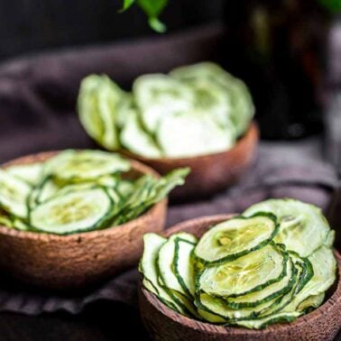 Three wooden bowls filled with thinly sliced cucumber chips rest on a dark cloth, offering fresh and delicious snack ideas against a blurred background.