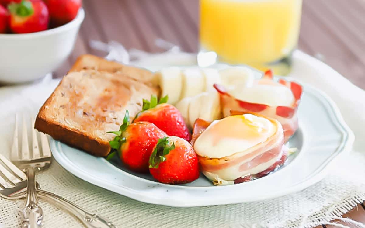 A plate with toast, strawberries, pear slices, bacon-wrapped egg, and apple slices with cheese—this healthy breakfast pairs perfectly with a glass of orange juice and a bowl of strawberries in the background.