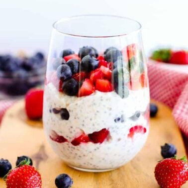 A glass filled with chia seed pudding layered with fresh strawberries and blueberries, placed on a wooden board with scattered berries and a red napkin—perfect for those seeking easy breakfasts or healthy breakfast ideas.