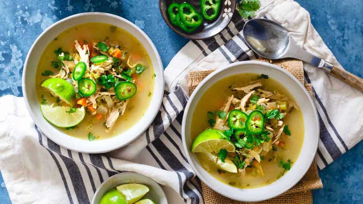 Two bowls of chicken soup, a classic in healthy soups, are garnished with lime wedges, cilantro, and sliced jalapeños, placed on a striped towel with a spoon and bowls of lime and jalapeño on the side. Perfect for fans of comforting soup recipes.