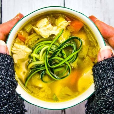 A person holding a bowl of chicken soup with vegetables and spiralized zucchini on top, wearing a dark knitted sweater—perfect for enjoying the warming recipes and soup benefits on a chilly day.