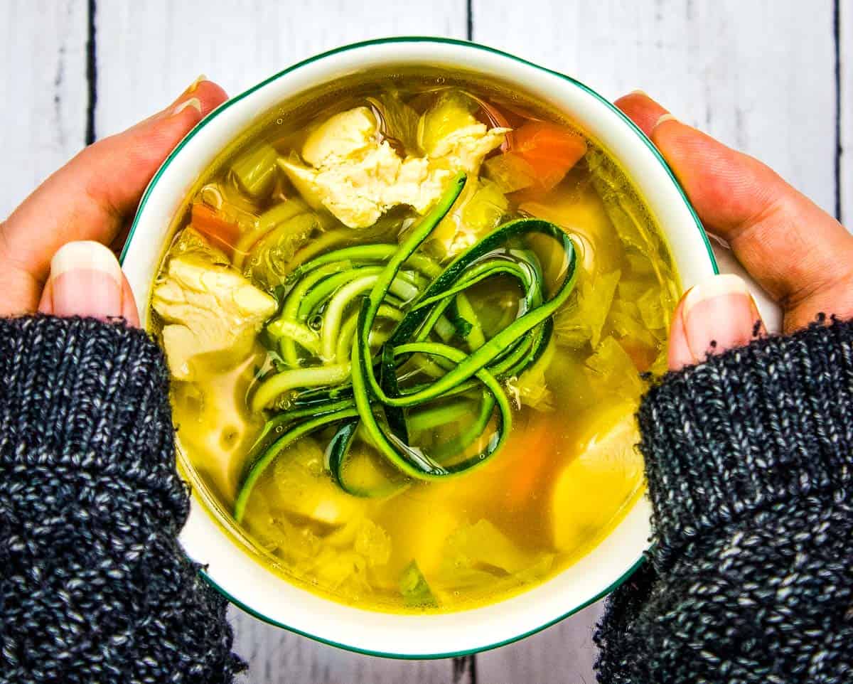 A person holding a bowl of chicken soup with vegetables and spiralized zucchini on top, wearing a dark knitted sweater—perfect for enjoying the warming recipes and soup benefits on a chilly day.