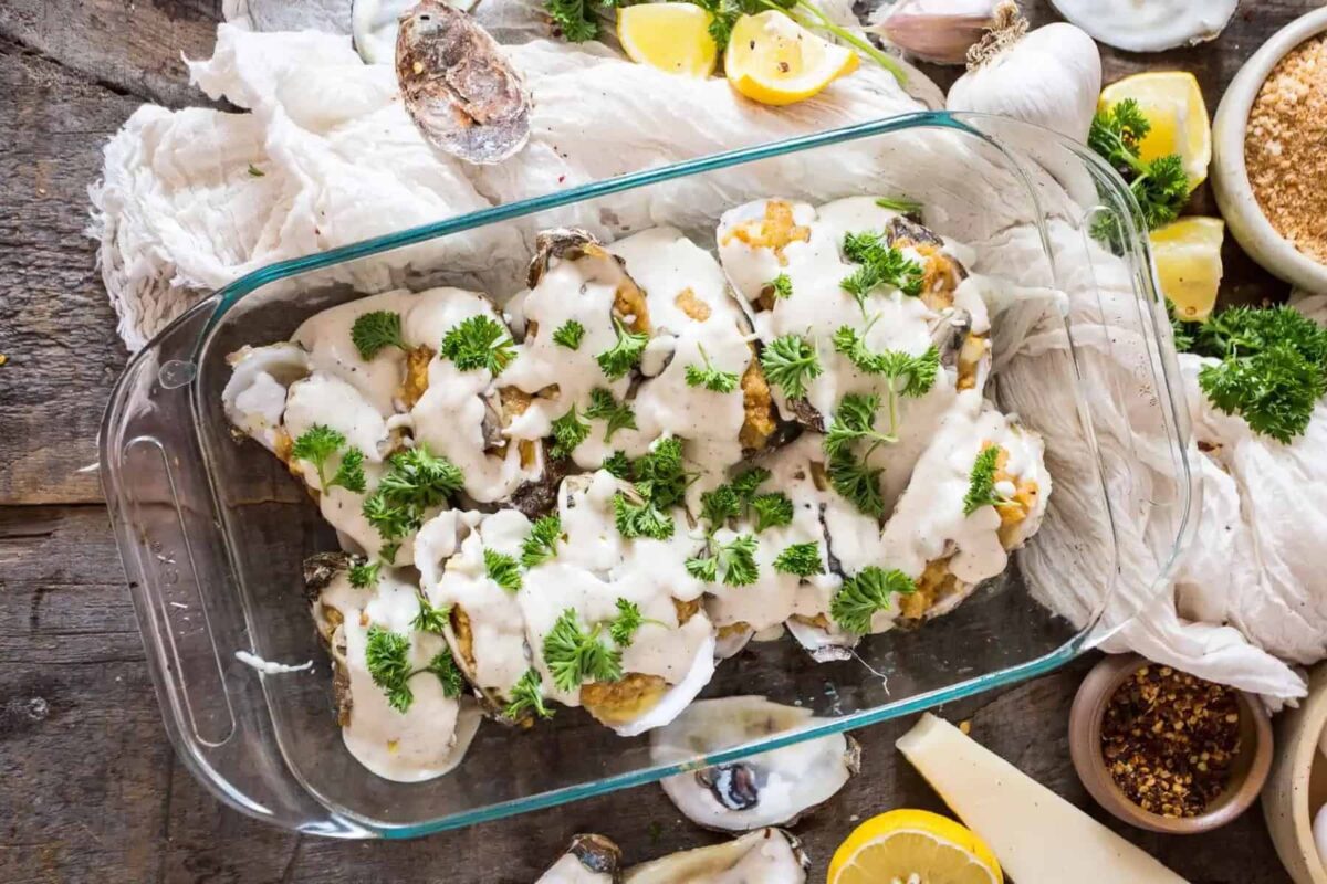 A glass baking dish filled with baked oysters topped with creamy white sauce and fresh parsley showcases classic ocean flavors, surrounded by lemon wedges, parsley, and various ingredients on a rustic wooden table.