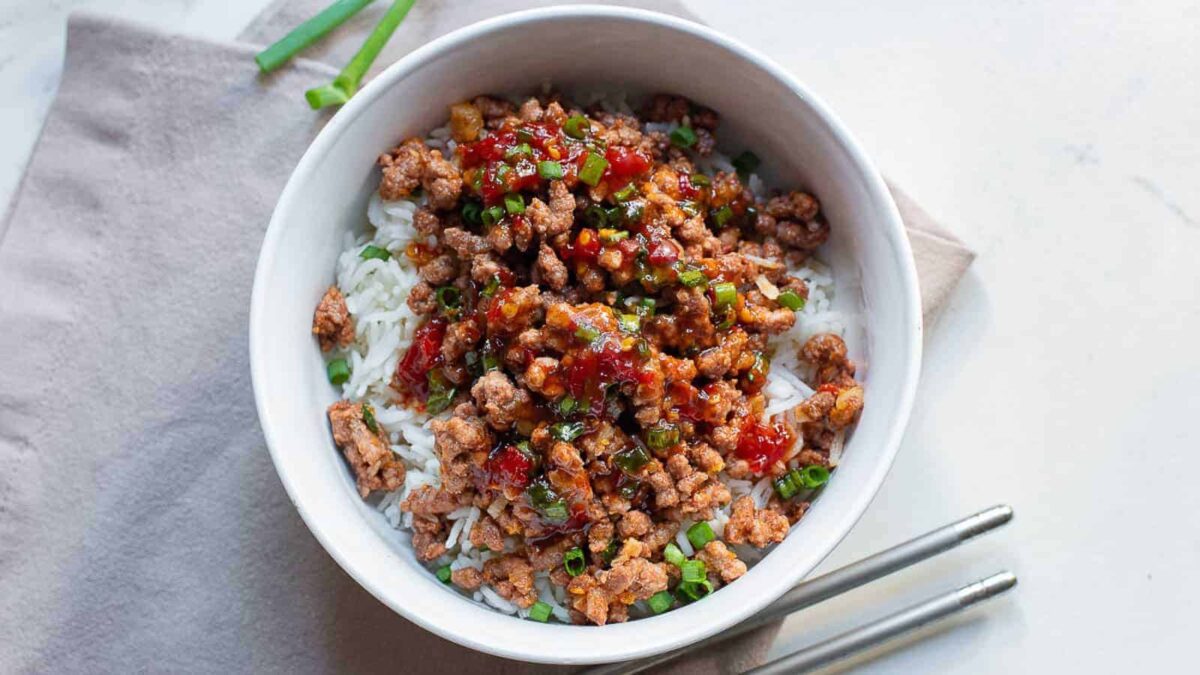 A bowl of white rice topped with seasoned ground beef, chopped scallions, and a red sauce, with chopsticks placed beside the bowl—perfect for those seeking easy beef meals or quick beef dinners.