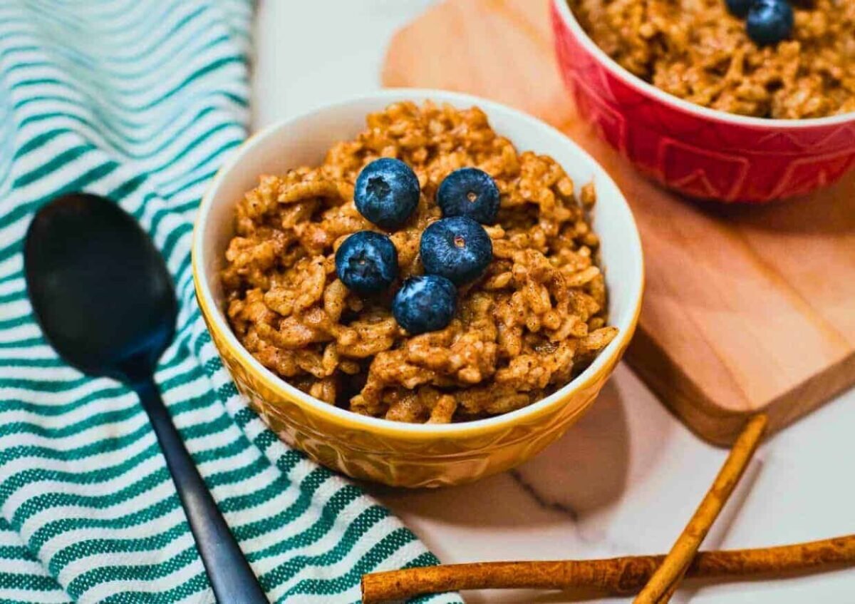 A bowl of rice pudding topped with fresh blueberries sits on a striped cloth beside a spoon, with another bowl and cinnamon sticks nearby—a delightful example of fresh vegan dishes.