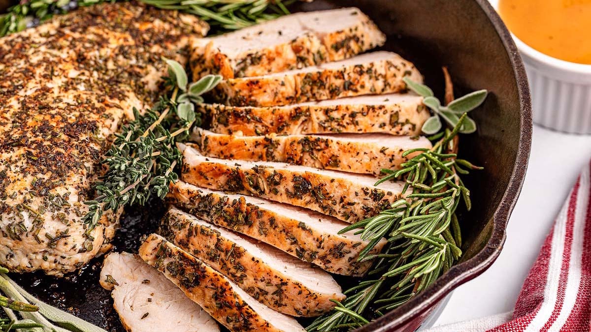 Herb-crusted turkey breast, sliced and arranged in a skillet with fresh rosemary and thyme, offers a flavorful twist on classic turkey dinners—perfect for delicious weeknight meals. Served with a small bowl of savory sauce.