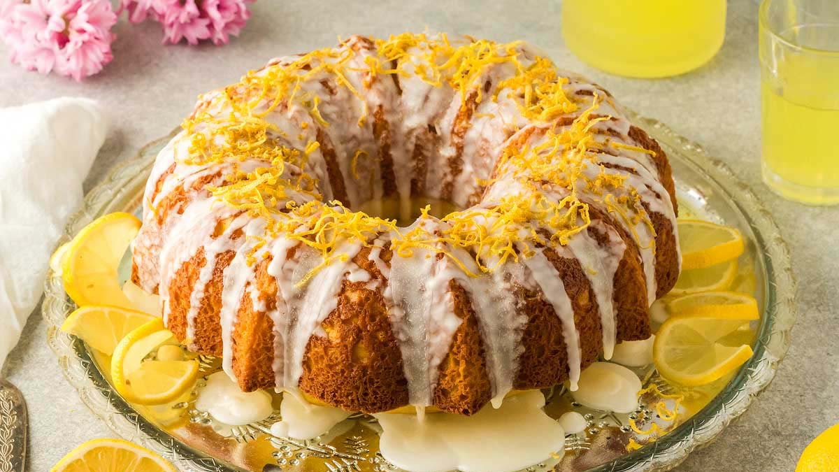 A must-try lemon bundt cake with white icing and lemon zest, beautifully served on a glass platter with lemon slices, alongside a refreshing lemon drink—perfect for Bundt cakes lovers.