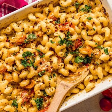A baked pasta casserole with cavatappi noodles, herbs, tomato pieces, and creamy feta in a white dish—one of those bold dishes perfect for trying new recipes. A wooden spoon and red napkin sit nearby.