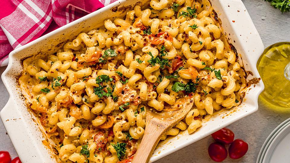 A baked pasta casserole with cavatappi noodles, herbs, tomato pieces, and creamy feta in a white dish—one of those bold dishes perfect for trying new recipes. A wooden spoon and red napkin sit nearby.