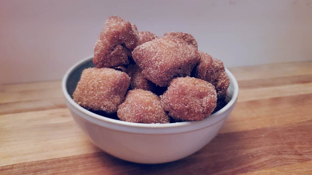 A white bowl filled with fluffy, sugar-coated doughnut bites sits on a wooden surface against a plain background.