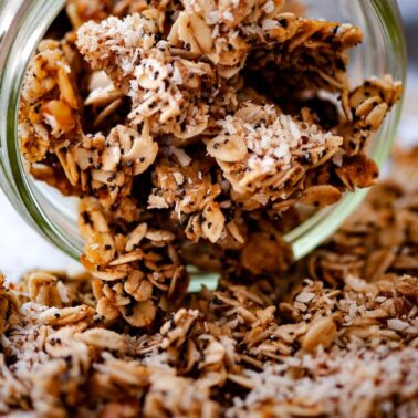 Loose granola with oats and coconut flakes spills from a glass jar onto a surface, perfect for topping your favorite fresh vegan recipes.